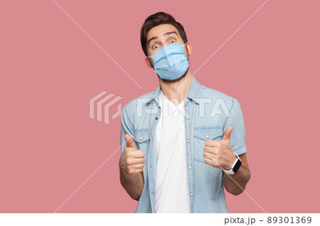 Portrait of young man with surgical medical mask in blue casual style shirt standing, thumbs up and looking at camera with surprised happy face. indoor studio shot, isolated on pink background. 89301369