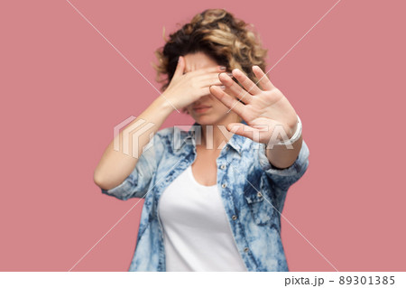 Stop, I don't want to look at this. Portrait of confused or scared young woman with curly hair standing covering her eyes and showing stop sign. indoor studio shot, isolated on pink background. 89301385