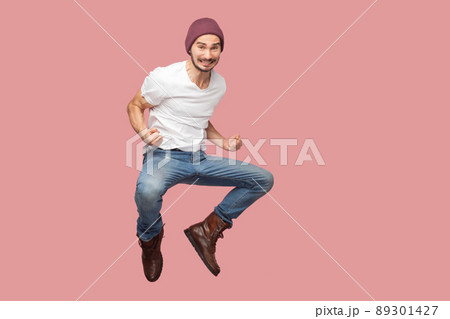 Portrait of happy bearded hipster young man in white shirt and blue jeans with hat jumping, rejoicing and celebrating his victory. indoor studio shot, isolated on pink background copyspace. Portrait of happy bearded hipster young man in white shirt and blue jeans with hat jumping, rejoicing and celebrating his victory. indoor studio shot, isolated on pink background copyspace. 89301427