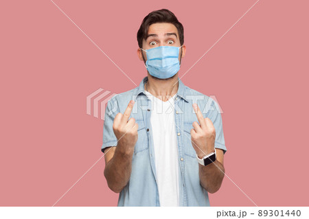 Portrait of angry young man with surgical medical mask in blue shirt standing with middle finger fuck sign hand gesture and looking at camera. indoor studio shot, isolated on pink background. 89301440