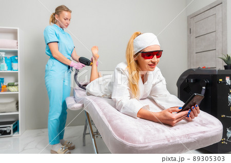 Beautician removes hair from the female's legs. Woman client lies on the couch and uses the phone during the procedure. The concept of laser hair removal in beauty salon Beautician removes hair from the female's legs. Woman client lies on the couch and uses the phone during the procedure. The concept of laser hair removal in beauty salon 89305303