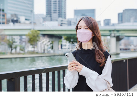 川沿いの遊歩道を地図アプリを見ながら歩く女 89307111