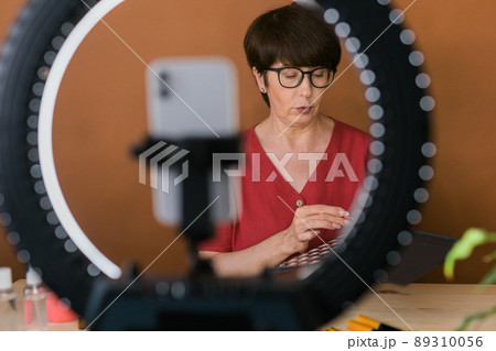 Middle-aged female beauty blogger or influencer with makeup product recording video in room at home. Using ring lamp and smartphone Middle-aged female beauty blogger or influencer with makeup product recording video in room at home. Using ring lamp and smartphone 89310056