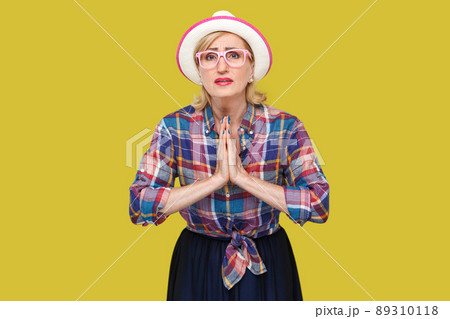 Please forgive me. or give me one more chance, Portrait of hopeful mature woman in casual style with hat and eyeglasses standing with palm hands and looking at camera, begging and asking. studio shot. 89310118