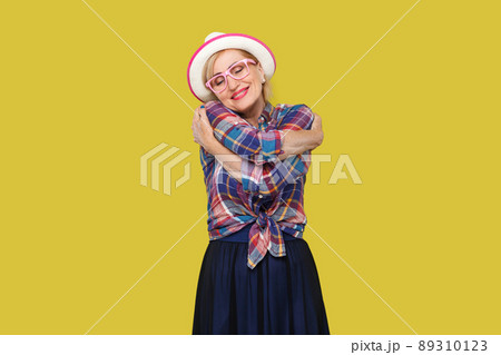 Portrait of peacful happy modern stylish mature woman in casual style with hat, eyeglasses standing, hugging herself with closed eyes and toothy smile. indoor studio shot isolated on yellow background 89310123