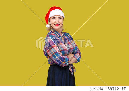 Portrait of confident attractive adult woman in red santa cap and checkered shirt standing and closed up hands with toothy smiling, looking at camera. Indoor, studio shot, yellow background 89310157