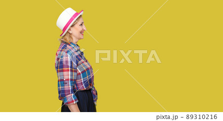 Profile side view portrait of happy successful modern stylish mature woman in casual style with hat standing, looking forward with toothy smile. indoor studio shot isolated on yellow background. Profile side view portrait of happy successful modern stylish mature woman in casual style with hat standing, looking forward with toothy smile. indoor studio shot isolated on yellow background. 89310216