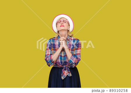 Oh my God please help me. Portrait of hopeful modern stylish mature woman in casual style with hat, eyeglasses standing with closed eyes and praying. indoor studio shot isolated on yellow background. Oh my God please help me. Portrait of hopeful modern stylish mature woman in casual style with hat, eyeglasses standing with closed eyes and praying. indoor studio shot isolated on yellow background. 89310258