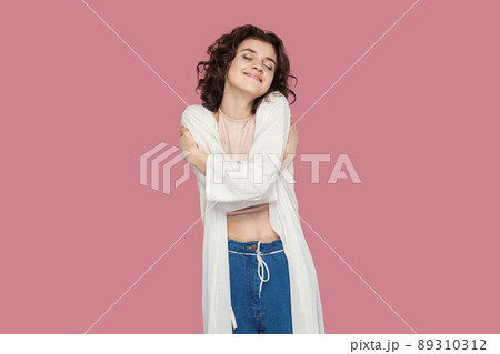 Portrait of happy satisfied beautiful brunette young woman with curly hairstyle in casual style standing, hugging herself and feeling happiness. indoor studio shot isolated on pink background. 89310312