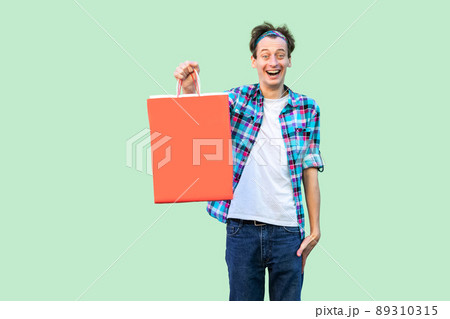Shocked modern young hipster man in white tshirt and checkered shirt standing and showing shopping bags and toothy smile, looking at camera. Studio shot, green background, isolated, indoor 89310315