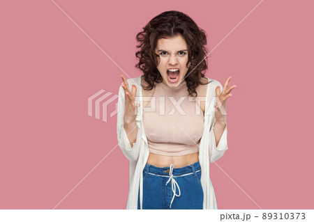 Portrait of angry brunette young woman with curly hairstyle in casual style standing, raised arms, screaming and looking at camera. expression and emotion. studio shot isolated on pink background. 89310373