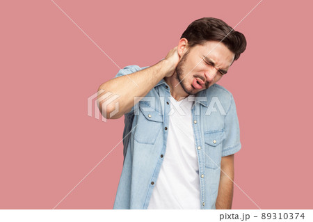 Neck or back pain. Portrait of sad sick handsome bearded young man in blue casual style shirt standing and holding his painful neck and feeling bad. indoor studio shot, isolated on pink background. 89310374