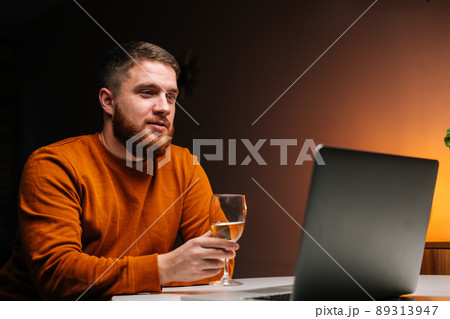 Close-up of young man enjoying distant dating using laptop, holding glass with wine, talking celebration toasting via video call, sitting at table with candle in dark room, looking on screen. Close-up of young man enjoying distant dating using laptop, holding glass with wine, talking celebration toasting via video call, sitting at table with candle in dark room, looking on screen. 89313947