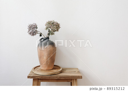 Elegant minimal summer, fall still life photo. Rustic clay vase, pitcher with hydrangea flowers on old wooden stool, console table. White wall background. Empty copy space. Elegant interior, lifestyle Elegant minimal summer, fall still life photo. Rustic clay vase, pitcher with hydrangea flowers on old wooden stool, console table. White wall background. Empty copy space. Elegant interior, lifestyle 89315212