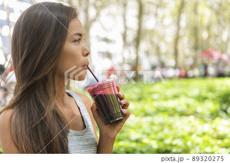 Woman drinking beet juice smoothie walking in city park. Girl eating detox food from online delivery service. Young asian biracial woman young adult living healthy lifestyle Woman drinking beet juice smoothie walking in city park. Girl eating detox food from online delivery service. Young asian biracial woman young adult living healthy lifestyle 89320275