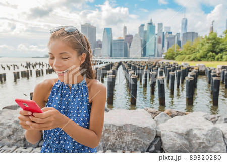 New York tourist woman using app on phone by Manhattan city skyline waterfront. People walking enjoying view of downtown from the Brooklyn Bridge park Pier 1 salt marsh New York tourist woman using app on phone by Manhattan city skyline waterfront. People walking enjoying view of downtown from the Brooklyn Bridge park Pier 1 salt marsh 89320280