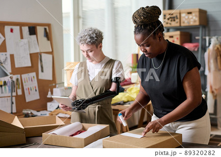Black woman sticking small paper with receiver address on packed box with goods while her colleague with disability using tablet 89320282