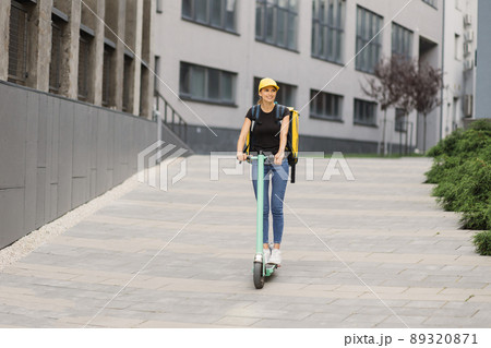 Delivery woman courier with isothermal backpack case rides her electric scooter 89320871