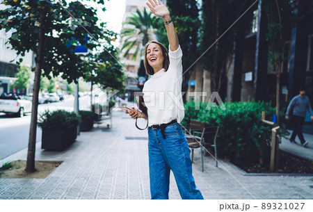 Happy young woman with smartphone waving hand on street 89321027