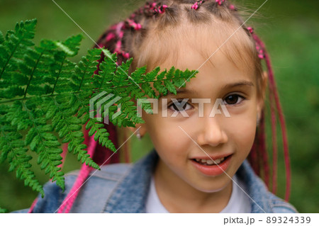 A little girl with African pink pigtails on a green natural background covers her face with a fern leaf. Cute beautiful child. Unity with nature. A little girl with African pink pigtails on a green natural background covers her face with a fern leaf. Cute beautiful child. Unity with nature. 89324339