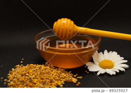 A yellow spoon of honey dipped in a jar of honey and bee bread scattered on a black background decorated with live chamomile. Honey dripping around, nice and inviting photos. Healthy food concept. A yellow spoon of honey dipped in a jar of honey and bee bread scattered on a black background decorated with live chamomile. Honey dripping around, nice and inviting photos. Healthy food concept. 89325027
