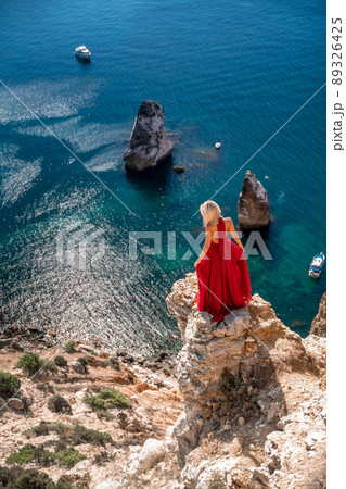 A girl with loose hair in a long red dress descends the stairs between the yellow rocks overlooking the sea. A rock can be seen in the sea. Sunny path on the sea from the rising sun 89326425