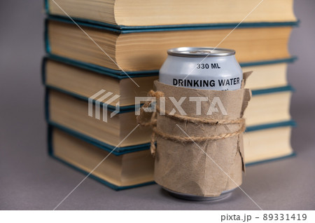 A stack of books and a can of drinking water against a gray background. Blue hardcover books. Can of freshwater wrapped in paper and tied with string. A stack of books and a can of drinking water against a gray background. Blue hardcover books. Can of freshwater wrapped in paper and tied with string. 89331419