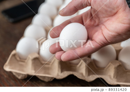 A man is holding a chicken egg in front of an egg tray. Adult male holding a white raw egg. Open recycled tray. Selective Focus. 89331420