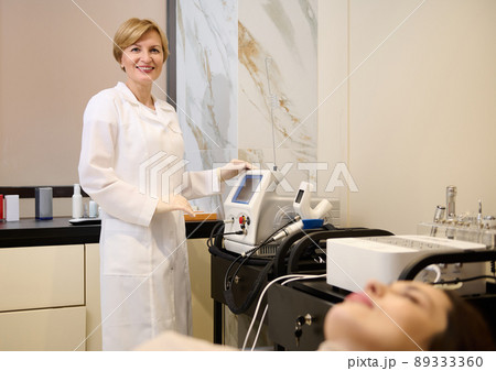Pleasant mature woman, female beautician standing near modern medical equipment for body beauty treatment in wellness spa clinic 89333360