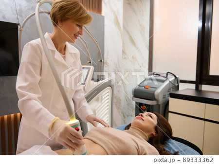 Pleasant aesthetician moving a radio frequency sensor over abdominal muscles of a pretty woman, for microstimulation, anti-cellulite treatment and weight loss in wellness spa Pleasant aesthetician moving a radio frequency sensor over abdominal muscles of a pretty woman, for microstimulation, anti-cellulite treatment and weight loss in wellness spa 89333362