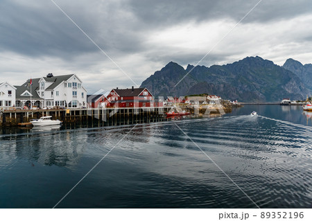 Norwegian seascape, cityscape of the town Henningsvaer, a small boat moves between peninsulas, sail boat, rocky coast with dramatic skies, classic view of Norwegian houses on the slopes Norwegian seascape, cityscape of the town Henningsvaer, a small boat moves between peninsulas, sail boat, rocky coast with dramatic skies, classic view of Norwegian houses on the slopes 89352196