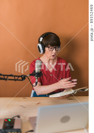 Middle-aged female radio presenter talking into the microphone and reading news - radio broadcast online concept Middle-aged female radio presenter talking into the microphone and reading news - radio broadcast online concept 89352469