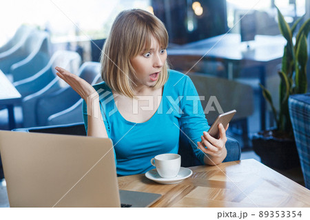 Top view portrait of young shocked blonde woman in blue t-shirt, sitting in cafe and holding her mobile smart phone and looking with amazed face and big eyes with raised hand. indoor studio shot. 89353354