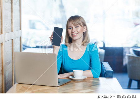 Portrait of happy beautiful young blonde woman in blue t-shirt, sitting with laptop, holding and showing mobile display and looking at camera with toothy smile. indoor, studio shot. 89353583