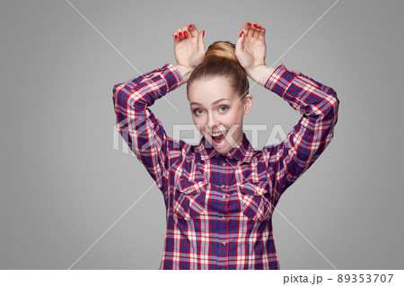 Funny beautiful blonde girl with pink checkered shirt, collected bun hairstyle and makeup standing and looking at camera with bunny gesture hands. indoor studio shot, isolated on gray background. Funny beautiful blonde girl with pink checkered shirt, collected bun hairstyle and makeup standing and looking at camera with bunny gesture hands. indoor studio shot, isolated on gray background. 89353707