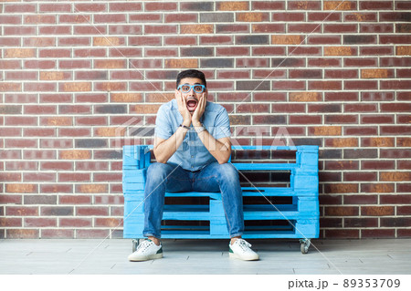 Full length portrait of amazed handsome young bearded man in casual style and eyeglasses sitting on blue wooden pallet and looking at camera with surprised face. indoor studio shot on brown brick wall Full length portrait of amazed handsome young bearded man in casual style and eyeglasses sitting on blue wooden pallet and looking at camera with surprised face. indoor studio shot on brown brick wall 89353709