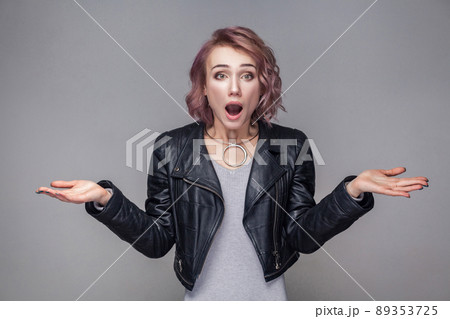 Portrait of shocked girl with short hairstyle, makeup in black leather jacket standing raised arms and looking at camera with big eyes and open mouth. indoor studio shot, isolated on grey background Portrait of shocked girl with short hairstyle, makeup in black leather jacket standing raised arms and looking at camera with big eyes and open mouth. indoor studio shot, isolated on grey background 89353725