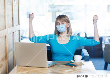 I am strong. Portrait of young woman with surgical medical mask in blue t-shirt is sitting and looking at camera and showing her biceps and satisfied. Indoor working, medicine and health care concept. 89353726
