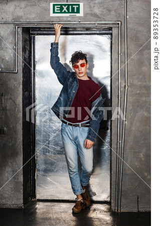 Full length portrait of stylish young fashion model man in bright red sunglasses and denim casual style posing near metallic door. standing and holding pipe. indoor, studio shot. Full length portrait of stylish young fashion model man in bright red sunglasses and denim casual style posing near metallic door. standing and holding pipe. indoor, studio shot. 89353728