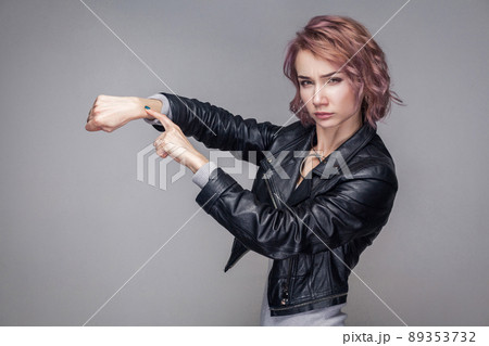 Portrait of serious beautiful girl with short hair and makeup in casual style black leather jacket standing, looking at camera and showing time gesture. indoor studio shot, isolated on grey background 89353732