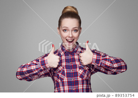 Satisfied beautiful girl with pink checkered shirt, collected bun hairstyle and makeup standing and showing thumbs up, looking at camera with amazed face. studio shot, isolated on gray background Satisfied beautiful girl with pink checkered shirt, collected bun hairstyle and makeup standing and showing thumbs up, looking at camera with amazed face. studio shot, isolated on gray background 89353754