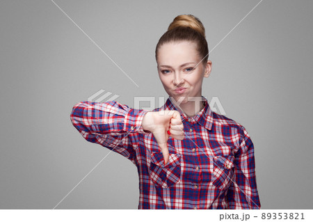 Unhappy sad girl with pink checkered shirt, collected bun hairstyle and makeup standing and looking at camera with unsatisfied face and thumbs down. indoor studio shot, isolated on gray background. Unhappy sad girl with pink checkered shirt, collected bun hairstyle and makeup standing and looking at camera with unsatisfied face and thumbs down. indoor studio shot, isolated on gray background. 89353821
