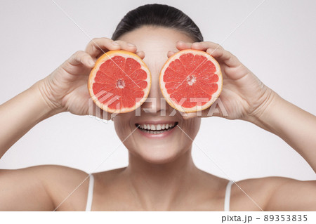 Closeup portrait of happy woman with perfect skin smiling after cream, balm, mask, lotion, holding half of grapefruit and covering her eye, isolated on grey background. Indoor, studio shot,copy space 89353835