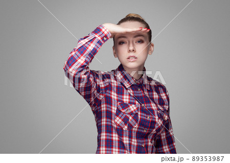 portrait of amazed beautiful blonde girl in pink checkered shirt, collected bun hairstyle and standing and looking at camera with hand on head and open mouth. studio shot, isolated on gray background 89353987