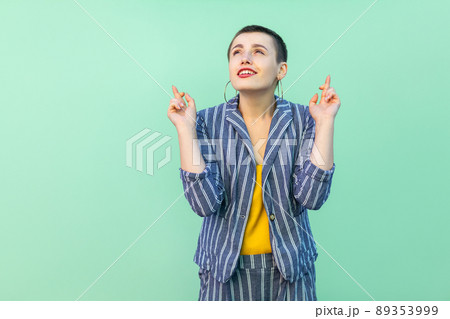 Portrait of hopeful handsome beautiful short hair young stylish woman in casual striped suit standing, crossed fingers and wishful to win . indoor studio shot isolated on light green background. Portrait of hopeful handsome beautiful short hair young stylish woman in casual striped suit standing, crossed fingers and wishful to win . indoor studio shot isolated on light green background. 89353999