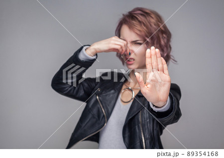 Stop, bad smell. Portrait ignoring of beautiful girl with short hairstyle and makeup in casual style black leather jacket standing pinching her nose. indoor studio shot, isolated on grey background. Stop, bad smell. Portrait ignoring of beautiful girl with short hairstyle and makeup in casual style black leather jacket standing pinching her nose. indoor studio shot, isolated on grey background. 89354168