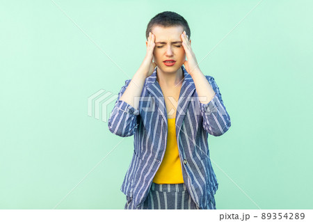 Headache or confusion. Portrait of handsome beautiful short hair young woman in casual striped suit standing holding her painful head or thinking. studio shot isolated on light green background. Headache or confusion. Portrait of handsome beautiful short hair young woman in casual striped suit standing holding her painful head or thinking. studio shot isolated on light green background. 89354289