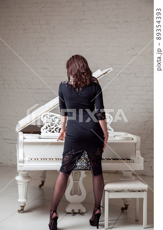 Back side view of seductive woman in dark classic dress playing piano and posing. indoor studio shot on white brick wall. Back side view of seductive woman in dark classic dress playing piano and posing. indoor studio shot on white brick wall. 89354393