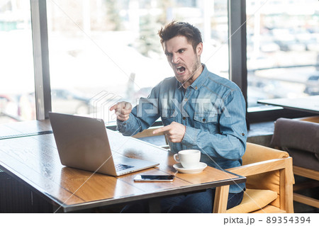 Yes, it`s you! Side view portrait of angry upset bearded young big boss in blue jeans shirt are sitting in cafe, working and making video call on laptop, pointing finger and blame the companion. Yes, it`s you! Side view portrait of angry upset bearded young big boss in blue jeans shirt are sitting in cafe, working and making video call on laptop, pointing finger and blame the companion. 89354394