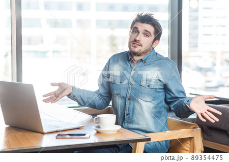 Not sure! Portrait of confused handsome bearded young freelancer in blue jeans shirt are sitting in cafe and working on laptop with raised arms and puzzled face, looking at camera. indoor 89354475